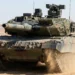 A front-view of the German Leopard 2A7+ Main Battle Tank (MBT) maneuvering in desert terrain. The image highlights the Rheinmetall 120mm L55A1 smoothbore gun, modular add-on armor for urban protection, and the FLW 200 remote weapon station on the turret.