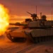 A US Army M1A2 SEPv3 Abrams tank in tan desert camouflage firing its 120mm main gun during a sunset exercise. A massive muzzle flash and dust cloud are visible. The tank's turret is fitted with the Trophy Active Protection System radar panels and countermeasure launchers. Tracks are kicking up sand as the tank moves.