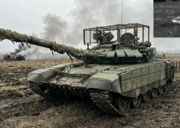 A Russian T-90M Proryv-3 Main Battle Tank maneuvering through muddy terrain in Ukraine. The tank is equipped with Relikt Explosive Reactive Armor (ERA) and covered in the Nakidka radar-absorbent camouflage system to reduce thermal signature against drones.