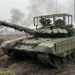 A Russian T-90M Proryv-3 Main Battle Tank maneuvering through muddy terrain in Ukraine. The tank is equipped with Relikt Explosive Reactive Armor (ERA) and covered in the Nakidka radar-absorbent camouflage system to reduce thermal signature against drones.